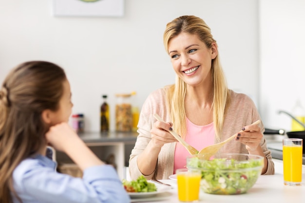 gezond eten, familie en mensenconcept - gelukkige moeder en dochter met groentesalade voor het avondeten in de keuken thuis