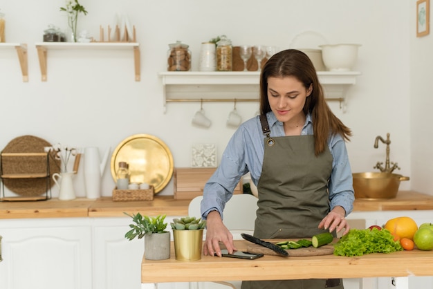 Gezond eten in huis. gelukkige jonge vrouw die de telefoon gebruikt om groentesalade in de keuken te bereiden.