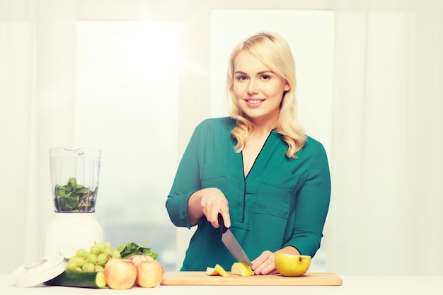 Foto gezond eten, koken, vegetarisch eten, dieet en mensenconcept - lachende jonge vrouw met blender en mes die groenten en fruit hakt op snijplank thuis