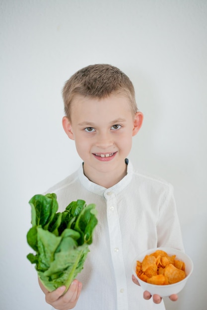 Gezond eten Schattige kleine jongen van 7 jaar oud houdt in de ene hand groene slablaadjes in de andere hand bord met vette frietjes House Eco