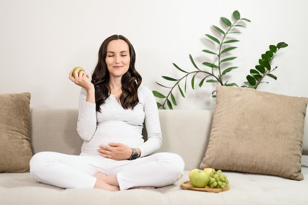 Gezond eten tijdens de zwangerschap, zwangere blanke vrouw eet groene appels zittend op de bank, gezonde snack met fruit