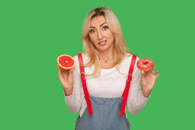 Gezond eten vs junkfood portret van verwarde vrouw met donut en verse grapefruit op zoek met twijfels kiezen tussen zoete bakkerij en fruit studio opname geïsoleerd op groene achtergrond