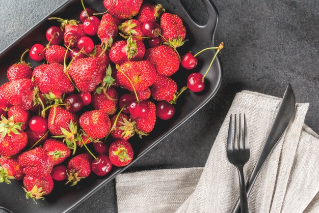 Gezond ontbijt, lunch of snack. Zomerbessen en fruit. Biologische verse kersen en aardbeien op een rechthoekige schotel, met een vork, op een zwarte stenen tafel. Bovenaanzicht