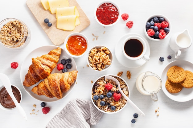 Gezond ontbijt met muesli, bessen, noten, croissants, jam, chocopasta en koffie. Bovenaanzicht