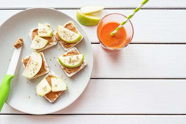 Gezond ontbijt met pindakaas en appel sandwiches op rijstwafels wortelsap Bovenaanzicht op witte houten achtergrond kopie ruimte