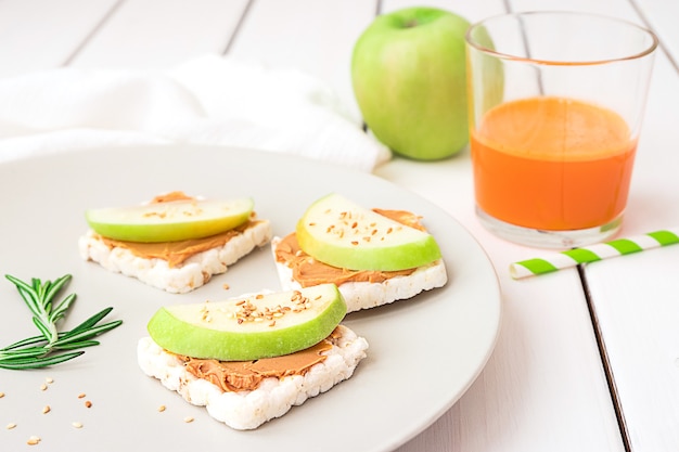 Gezond ontbijt met pindakaas en appelsandwiches op rijstwortels, wortelsap en appel op witte houten achtergrond hoogste mening