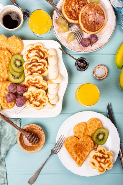 Gezond ontbijt met verse warme wafels harten, pannenkoeken bloemen met bessenjam en fruit op turquoise oppervlak, bovenaanzicht, plat leggen. Voedsel concept