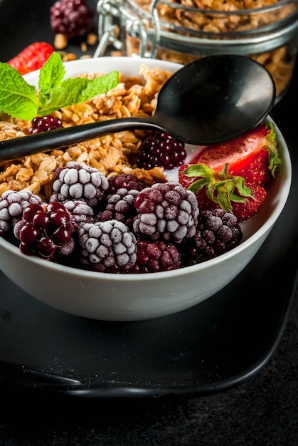 Foto gezond ontbijt. zomerbessen en fruit. zelfgemaakte griekse yoghurt met granola, bramen, aardbeien en munt. zwarte stenen tafel, met de ingrediënten.