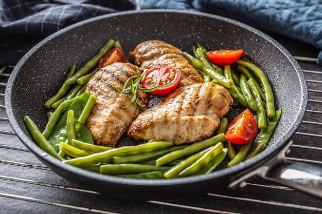 Gezond recept van gegrilde kip, groene groente en tomaten op een donkere pan met antiaanbaklaag op een metalen rooster en een donkere achtergrond.