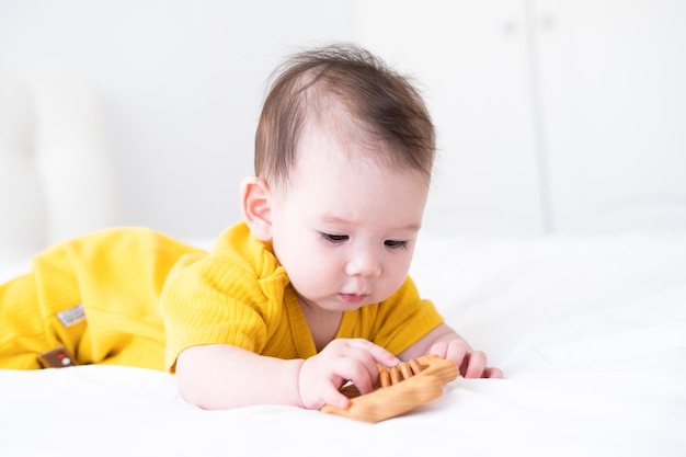 Gezonde aziatische babymeisje in gele bodysuit speelt met houten speelgoed bijtring voor tanden op wit beddengoed