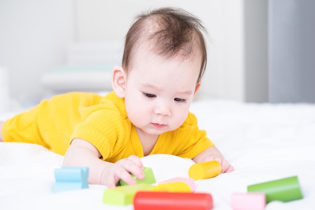 Gezonde aziatische babymeisje in gele bodysuit spelen met houten speelgoed op wit beddengoed