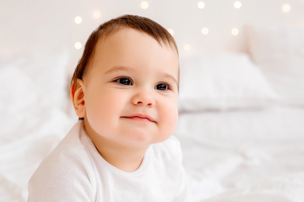Gezonde babyjongen 10 maanden oud in witte kleren glimlachend zittend op wit beddengoed in bed, ruimte voor tekst