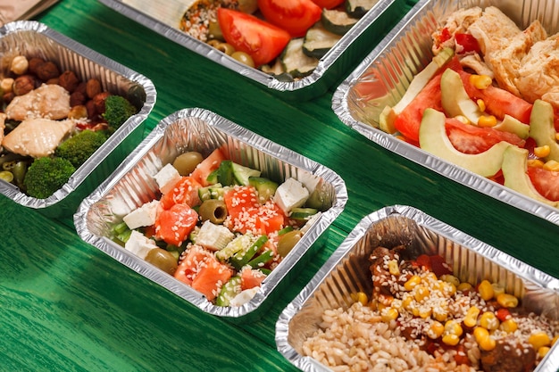 Gezonde bezorging van restaurantmaaltijden. Take away van dagelijkse maaltijden in folie lunchboxen met kopieerruimte bij groen hout. Salades, vlees en fruit.