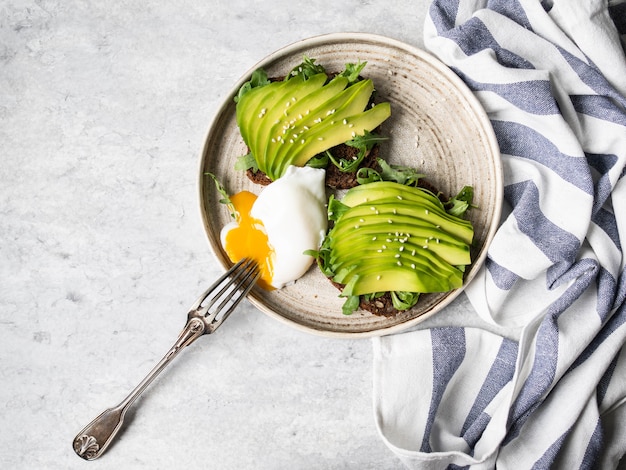 Gezonde eigengemaakte toosts met rucola en avocado op het brood van de roggekorrel en gepocheerd ei met het uitspreiden van dooier op plaat op grijze achtergrond. Bovenaanzicht