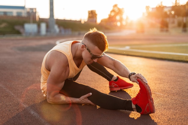 Gezonde fitness man atleet met stijlvolle zonnebril in mode sportkleding met rode hardloopschoenen doen een stuk en bereidt zich voor op de vlucht bij zonsondergang