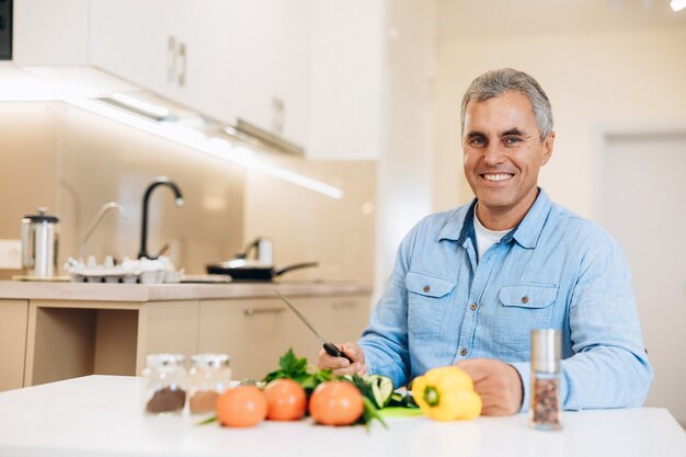 Gezonde geest in een gezond lichaam! Glimlachende volwassen man bereidt zich voor om de groenten te hakken om een veganistisch gerecht te koken. Gezond ouder worden concept. Moderne en lichte keuken interieur.