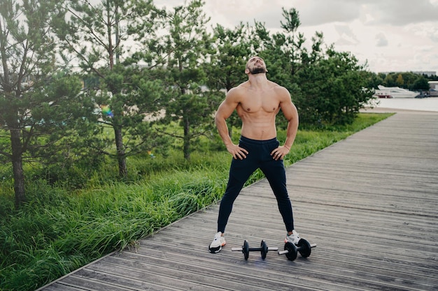 Gezonde gespierde man staat met naakte torso steekt het hoofd boven en ademt diep draagt sportkleding heeft training met halters poses buiten Bodybuilding actieve levensstijl fitness concept