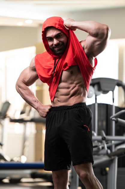 Gezonde jonge Man permanent sterk in de sportschool en buigen spieren in een rode Hoodie gespierde atletische Bodybuilder Fitness Model poseren na oefeningen