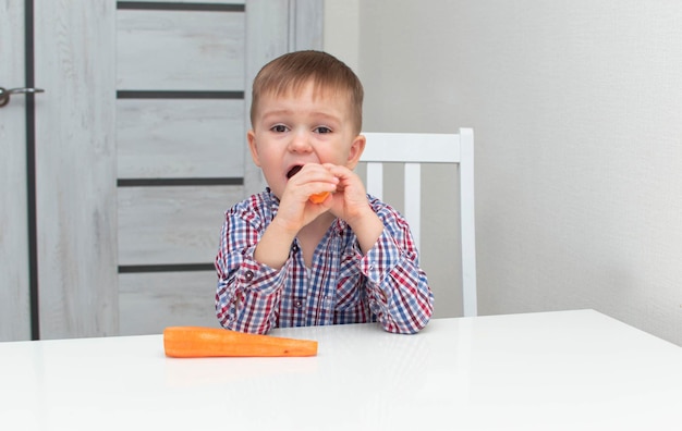 Gezonde jongen met grote bruine ogen zit aan witte tafel en eet een oranje wortel. Thuis gezond eten.