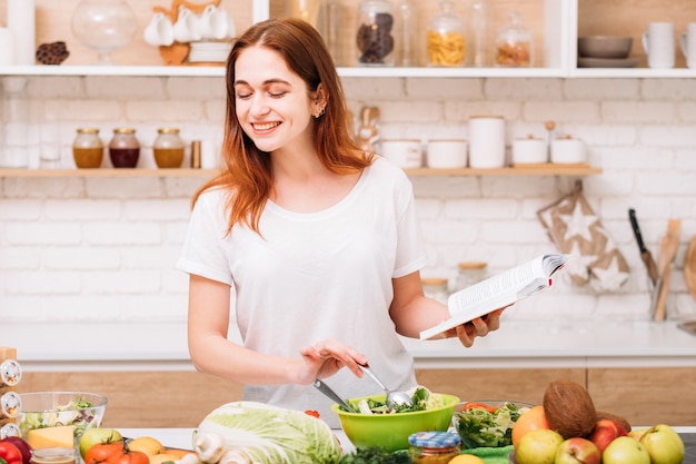 Gezonde levensstijl. Biologisch diner recept. Groen eten om fit te zijn. Glimlachende jonge vrouw die met boek in handen maaltijd voorbereiden.