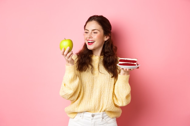 Gezonde levensstijl concept jonge brunette vrouw met groene appel vol vitaminen en cake met veel calorieën glimlachend en starend naar fruit staande tegen roze muur