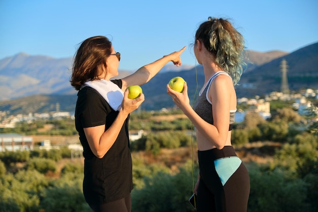 Gezonde levensstijl en gezond voedselconcept. Glimlachende fitnessmoeder en tienerdochter die samen appels eten, praten, rusten na atletisch hardlopen en buitensporten