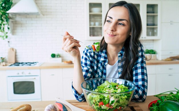 Gezonde levensstijl. Goede leven. Biologisch voedsel. Groenten. Close-up portret van een gelukkige, schattige, mooie jonge vrouw terwijl ze thuis smakelijke veganistische salade in de keuken probeert