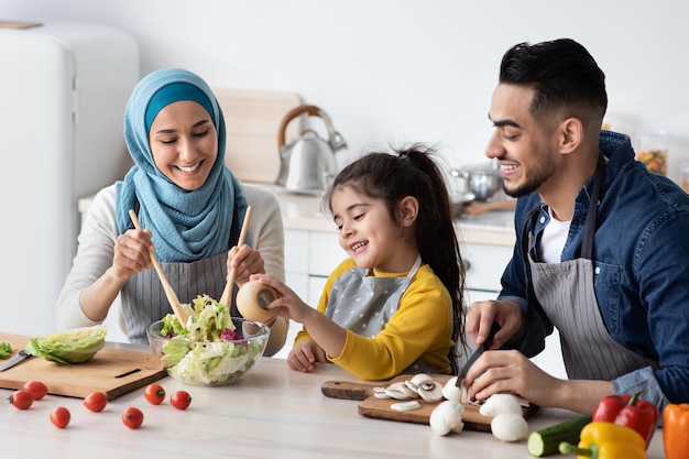 Gezonde lunch. vrolijke moslimgezin van drie koken samen vegetarische maaltijd in de keuken, gelukkige midden-oosterse ouders en hun dochtertje bereiden groentesalade, kruiden, close-up toe te voegen