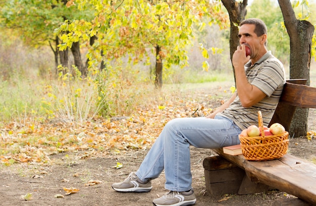 Gezonde man van middelbare leeftijd die alleen zit op een rustieke houten bank in het bos met een mandje met vers geplukte appels die een appel eten