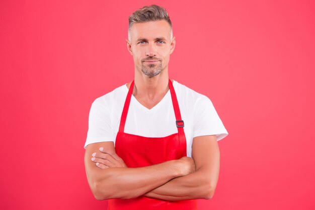 Gezonde manier koken. Meester van het huishouden. Huishouden doen. Thuis koken. Grillfeestje. Bekwaam en zelfverzekerd. Kok die slabschort draagt. Bebaarde volwassen man in rode schort. Volwassen man in kookschort.