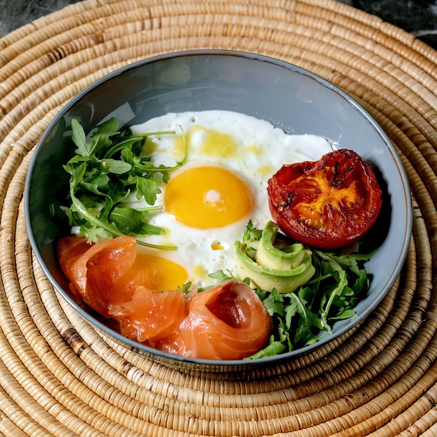 Gezonde ontbijtkom met gebakken eieren, zalm, avocado, gegrilde tomaat en salade serveren met brood op stro servet over donkere marmeren achtergrond. Vierkante afbeelding
