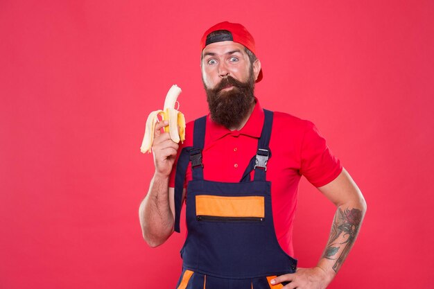 Gezonde pauze concept brute man in pet en uniform eet banaan gezonde lunch snack volwassen man bouwer voel honger mens kracht en gezondheid rechtopstaande banaan als concept van het behandelen van impotentie