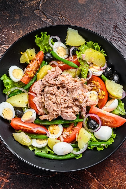 Gezonde stevige salade met tonijn, sperziebonen, tomaten, eieren, aardappelen en zwarte olijven in een bord. Donkere achtergrond.