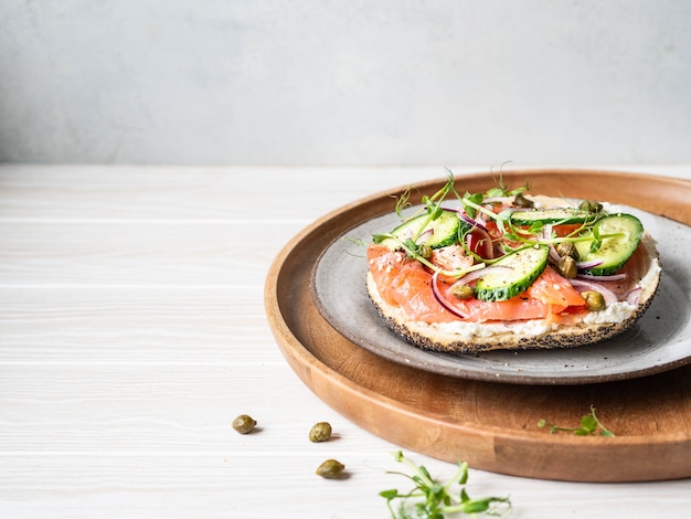 Gezonde toast met roomkaas, zalm, verse komkommer, kappertjes, rode ui, zwarte peper en erwtenspruiten op plaat.