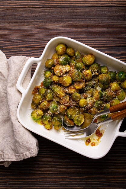 Gezonde vegetarische schotel geroosterde spruitjes met boter en parmezaanse kaas in witte keramische braadpan bovenaanzicht op donkere rustieke houten tafel van bovenaf, veganistisch eten