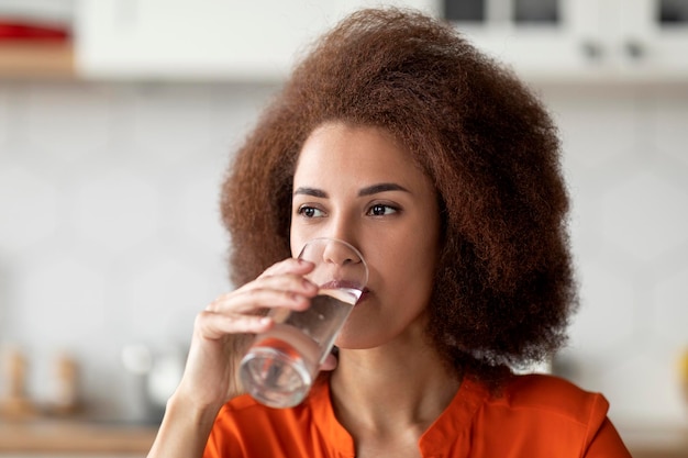 Gezonde vloeibare mooie zwarte vrouw die mineraalwater drinkt uit glas in de keuken