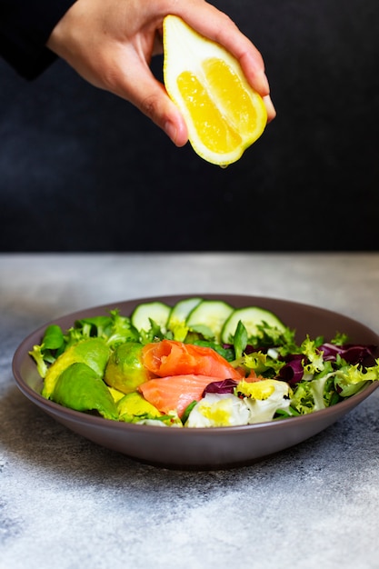 Gezonde voeding salade met zalm, avocado, pompoenpitten, verse groenten en citroen geserveerd op een grijze tafel. Vrouwelijke hand knijpt citroen op de salade. Het concept van gezond eten. Zwarte achtergrond