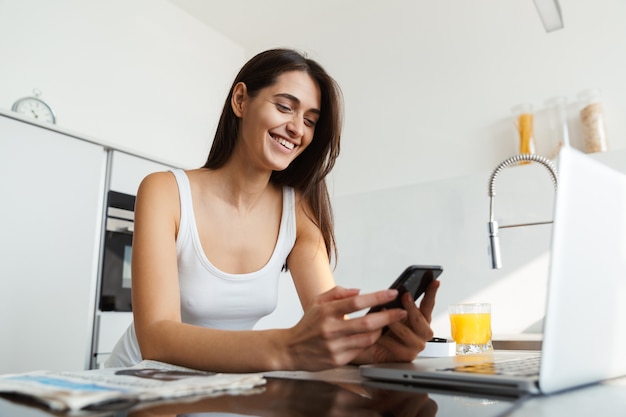 Gezonde vrouw in de keuken die zich dagelijkse ochtendroutine bevindt die door mobiele telefoon babbelt.