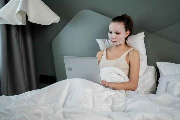 Gezonde vrouw zit op een bed met een cosmetisch gezichtsmasker met behulp van een laptop en werkt online hebben een