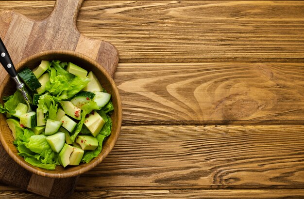 Gezonde zelfgemaakte avocado, komkommer, groene bladeren salade in houten kom op snijplank op houten rustieke bruine achtergrond. Vegetarisch eten, gezonde snack voor dieet en schoon eten. Bovenaanzicht, kopieer ruimte