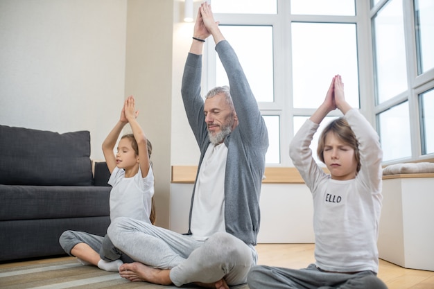 Gezondheid, yoga. volwassen grijsharige man en twee kinderen in de leerplichtige leeftijd kalm met gesloten ogen, opgeheven armen die thuis in yogahouding zitten