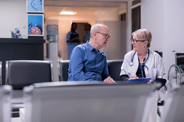 Gezondheidsspecialist in gesprek met senior patiënt, zittend in wachtruimte ziekenhuislobby, controleonderzoek doend bij medische afspraak. Medic en oude man bij overleg in wachtkamer, gezondheidszorg.