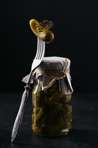 Gherkins pickled cucumber on a fork glass jar with marinated vegetables on a black background