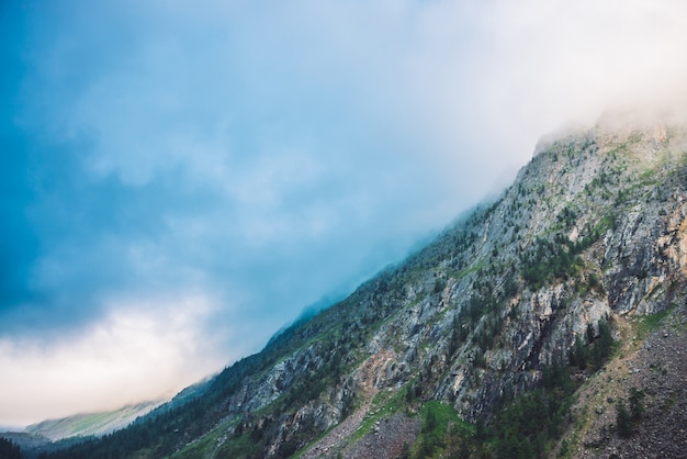 Giant cloud above rocky ridge with trees in sunlight. Amazing mountain range under blue cloudy dawn sky. Wonderful foggy rocks. Misty landscape of highland nature. Low clouds in morning sunny light.