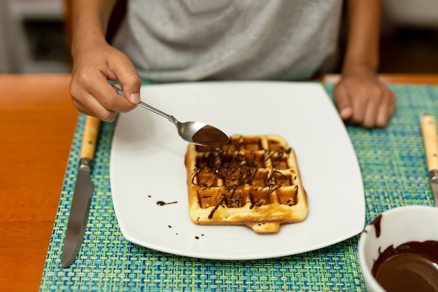 Foto giet de chocoladestroop van de kindhand op wafel in witte plaat bij ontbijt