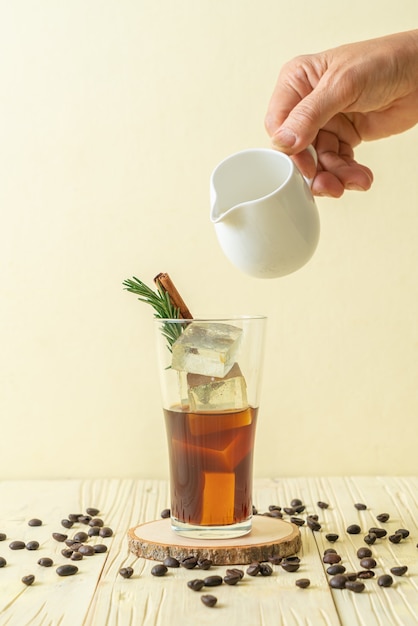 gieten melk in zwarte koffie glas met ijsblokje, kaneel en rozemarijn op houten oppervlak