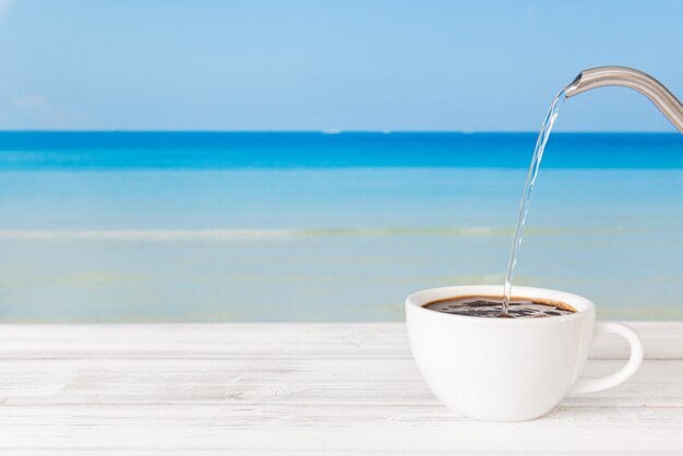 Gietend heet water in koffiekop op witte houten lijst met heldere blauwe overzeese achtergrond