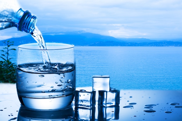 Gietend water van fles in een glas tegen de aardachtergrond met meer en plant dichtbij ijsblokjes