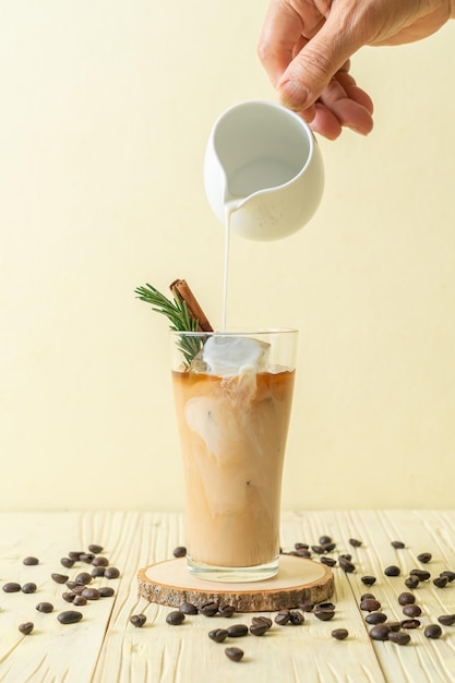 gietende melk in zwart koffieglas met ijsblokje, kaneel en rozemarijn op houten ondergrond