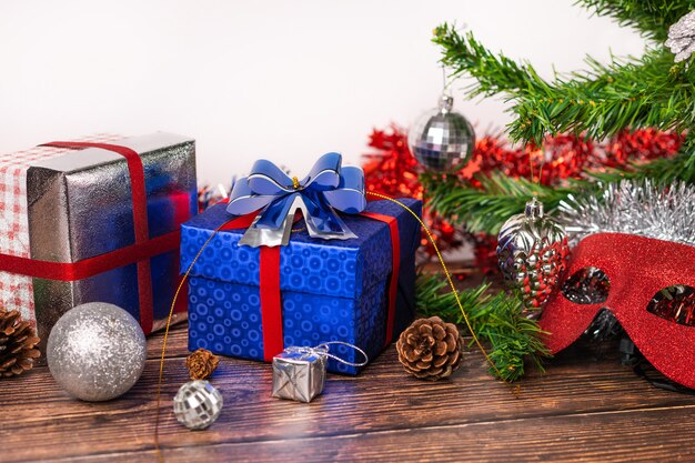 A gift in a blue wrapper, tied with a red ribbon, stands on a wooden table, with silver tinsel, Christmas balls and small decorative red mittens, a masquerade mask. Gifts.